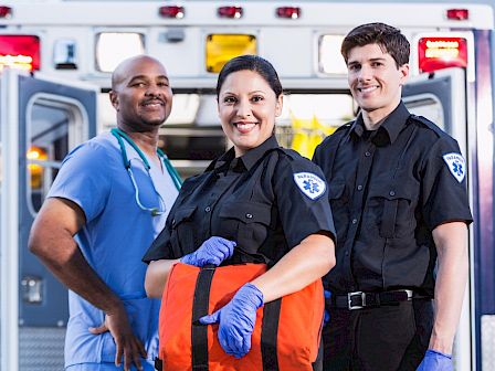 The image shows three medical professionals standing in front of an ambulance, including a doctor and two paramedics, all smiling at the camera.
