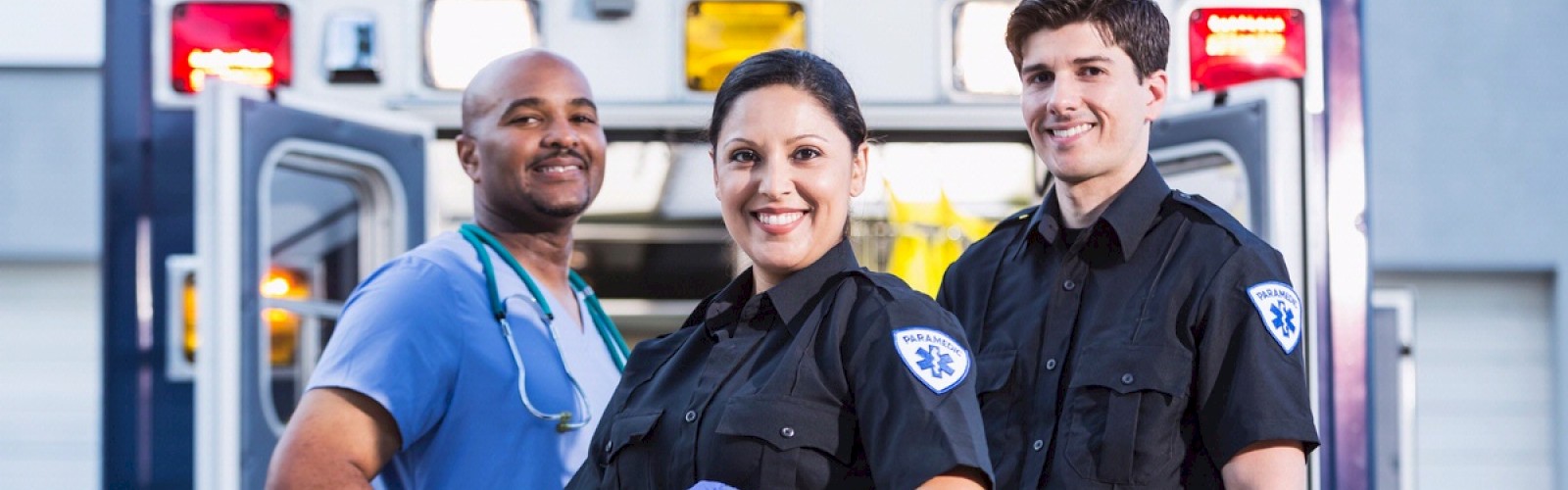 The image shows three medical professionals standing in front of an ambulance, smiling at the camera, with one wearing scrubs and two in EMS uniforms.