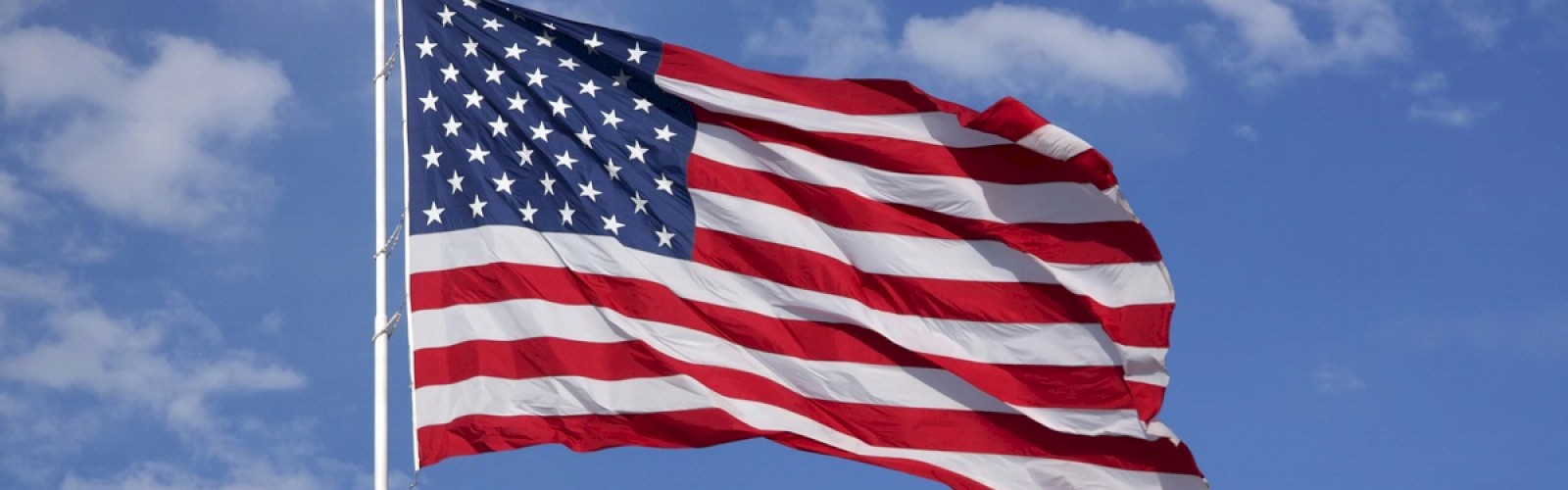 The image shows an American flag waving in the wind against a blue sky with scattered clouds.