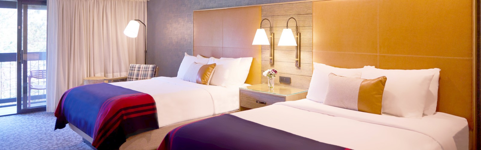 A Riverhouse Lodge hotel room with two neatly made queen-sized beds, matching lamps, a side table, blue accent wall, and a sliding glass door leading to a balcony.