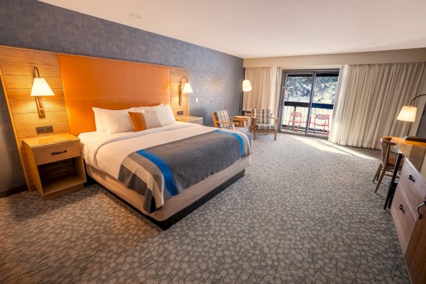 A cozy hotel room with a bed, nightstands, lamps, a chair, desk, curtains, and a sliding door leading to a balcony.