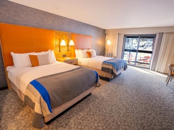 The image shows a hotel room with two double beds, bedside lamps, a desk, a chair, and a balcony with a view outside.