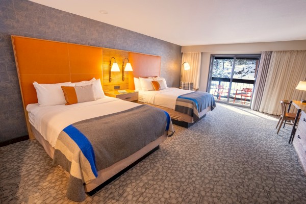 The image shows a hotel room with two double beds, bedside lamps, a desk, a chair, and a balcony with a view outside.