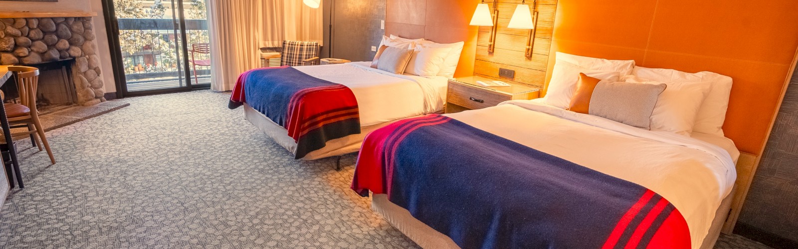 A cozy hotel room with two double beds, colorful bedding, warm lighting, a desk, and a view through the window.