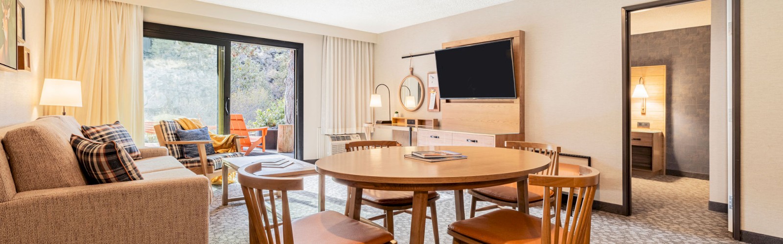 A cozy hotel suite at Riverhouse Lodge in Bend, Oregon with a living area, TV, sofa, table, chairs, and balcony view. A separate bedroom is visible through the open door.