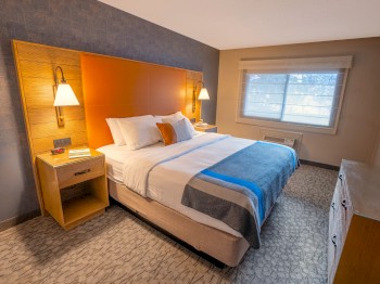 A cozy hotel room with a king-size bed, bedside tables with lamps, a dresser, carpeted floor, and a window covered with blinds.