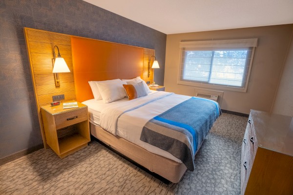 A cozy hotel room with a king-size bed, bedside tables with lamps, a dresser, carpeted floor, and a window covered with blinds.