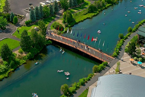 Aerial view of a scenic river with a bridge adorned with flags, surrounded by greenery. People are kayaking and exploring the water.