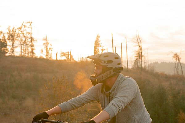 A person wearing a helmet and gloves is standing with their mountain bike in a scenic outdoor area during sunset.