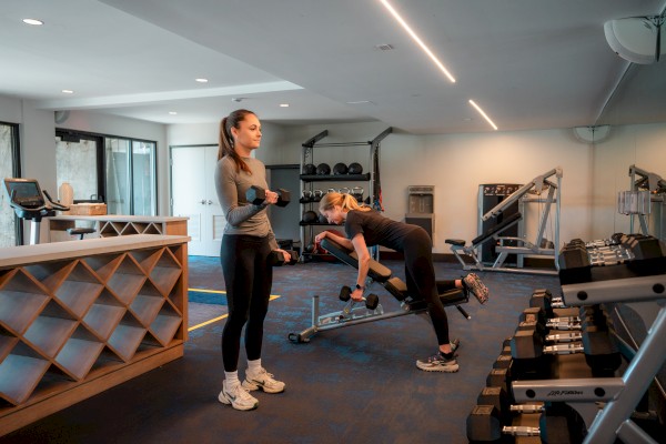 Two people are exercising in the gym at Riverhouse Lodge in Bend Oregon, using dumbbells and fitness equipment. The room is well-lit with various workout machines.