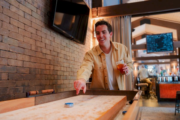A person is playing shuffleboard at Currents at Riverhouse Lodge in Bend, Oregon, holding a drink. They are in a casual setting with a TV and menu board in the background.