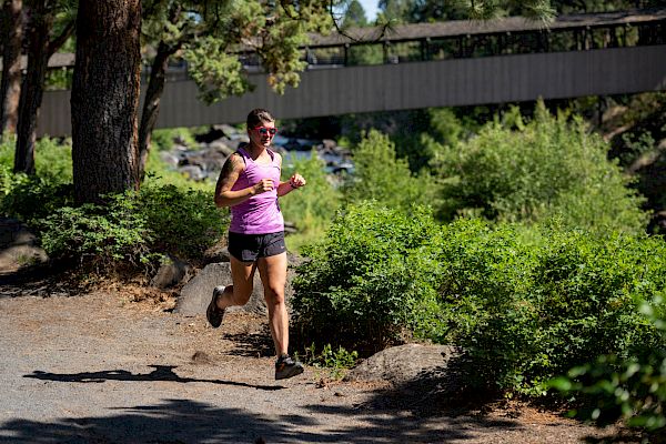 A person in athletic attire runs on a shaded trail with greenery and a bridge visible in the background, enjoying an outdoor workout.