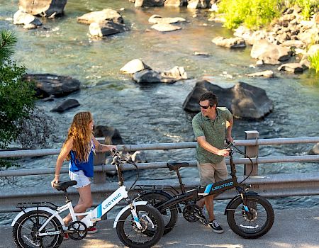 Two people are standing on a bridge with folding bikes, conversing with a scenic river and rocks in the background, enjoying the outdoors.