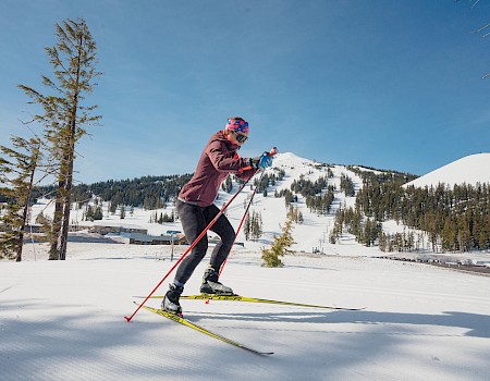 A person cross-country skiing in a snowy mountain landscape under a clear blue sky.