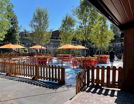 An outdoor seating area with wooden tables and orange umbrellas, surrounded by a wooden fence and trees on a sunny day.