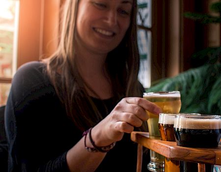 A woman is smiling and reaching for a glass of beer from a wooden tray that holds multiple glasses of different types of beer.
