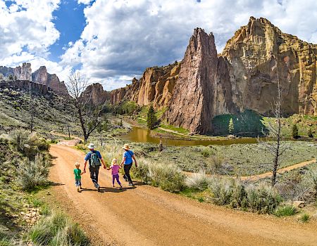 A family of four is walking on a dirt path surrounded by stunning rock formations and a clear sky with scattered clouds, enjoying a scenic hike.