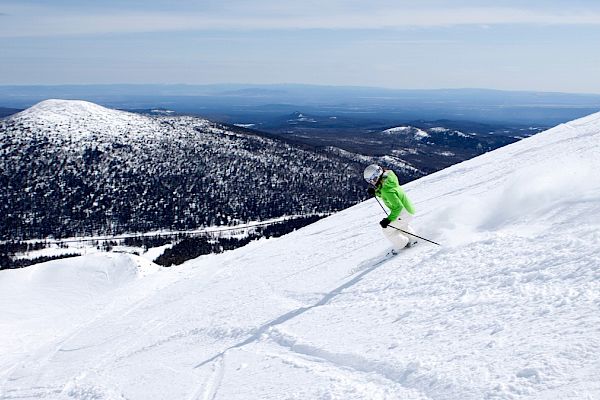 A skier in a green jacket is descending a snowy mountain with a scenic view of snow-covered hills and distant landscape in the background.