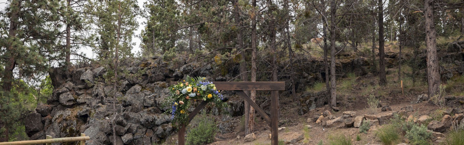An outdoor wedding setup with a wooden arch decorated with flowers and several black chairs, situated in a forested area with rocks and trees.
