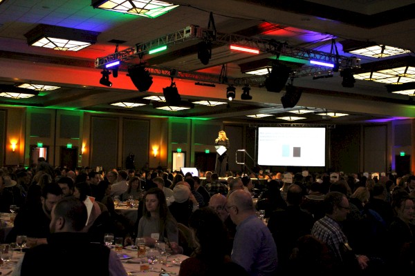 A large conference or event with many people seated at tables, and a speaker presenting on stage with a screen displaying a presentation.