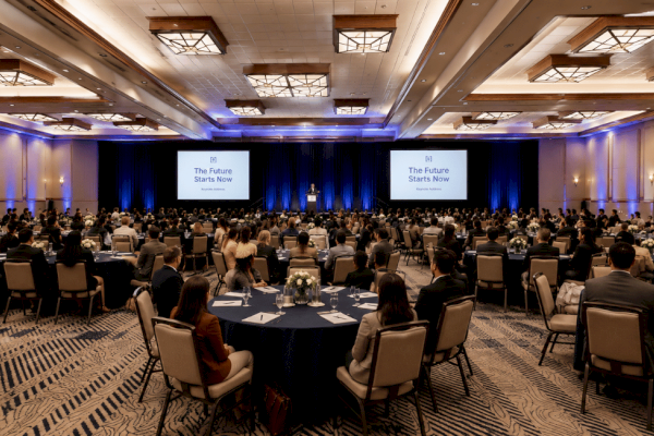 A conference with a speaker on stage at Riverhouse Lodge, multiple screens, and many attendees seated at round tables in a large, well-lit ballroom.