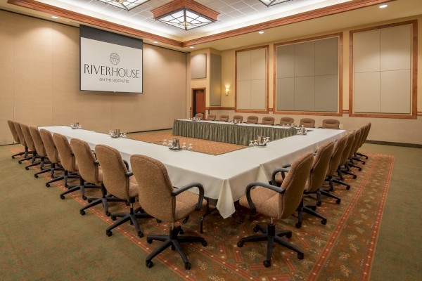 The image shows a conference room with a U-shaped table arrangement, chairs around the table, and a screen displaying 