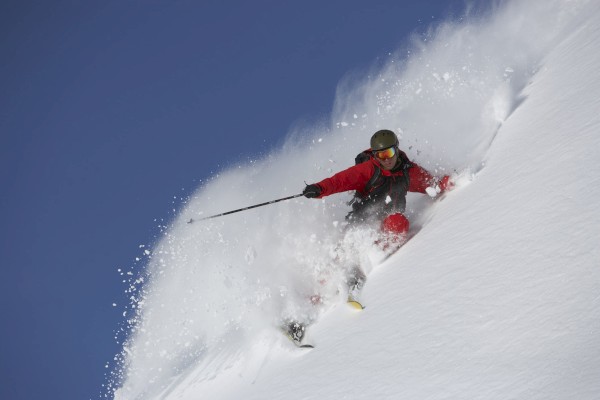A skier wearing a red jacket and helmet is descending a snowy slope, surrounded by a spray of powder.