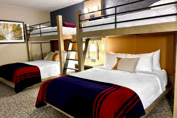 The image shows a hotel room with two queen beds, each has an overhead bunk bed and the room features modern decor and lighting.