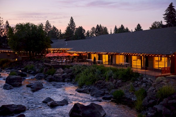 An outdoor restaurant with string lights is nestled by a rocky river and surrounded by trees at dusk, providing a cozy and scenic dining atmosphere.