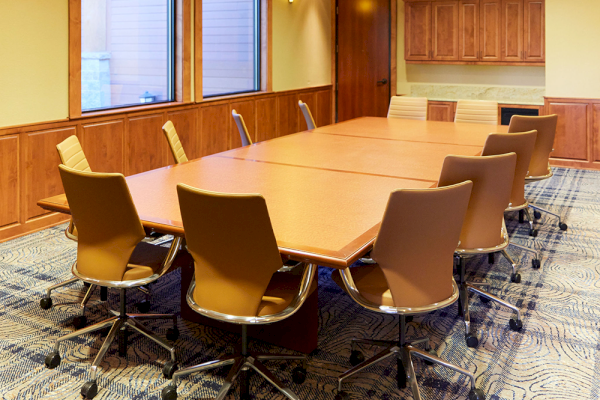A well-lit conference room with a long wooden table, eight chairs on wheels, wooden cabinets, and three windows letting in natural light.
