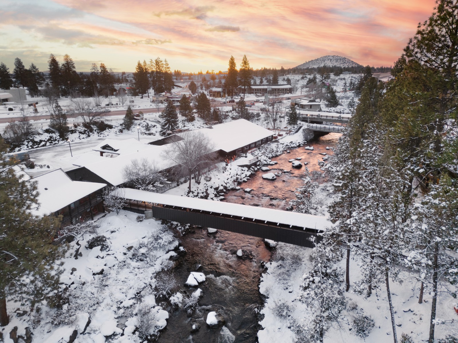 Snowy riverfront at Riverhouse Lodge in Bend, Oregon, with a covered bridge and buildings glowing under a colorful sunset sky.