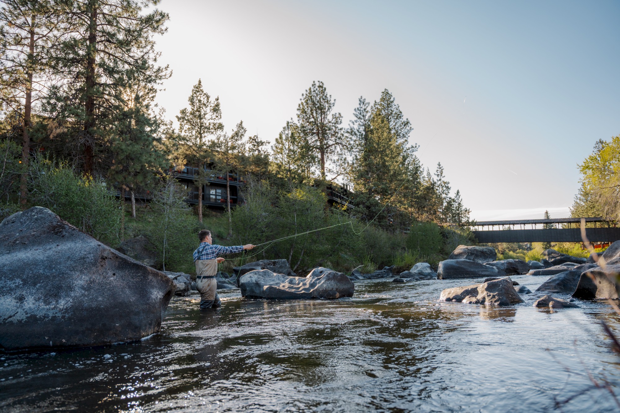 A man fly fishing in the Deschutes River in Bend, Oregon, with Riverhouse Lodge nestled among pine trees in the background and a wooden pedestrian bridge spanning the river.