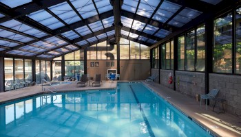 The image shows an indoor swimming pool with a glass ceiling and surrounding windows. There are chairs and a pool lift on the deck.