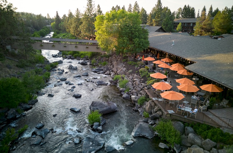 A riverside scene featuring rocky water, a bridge, a building with outdoor seating, and orange umbrellas, surrounded by trees.