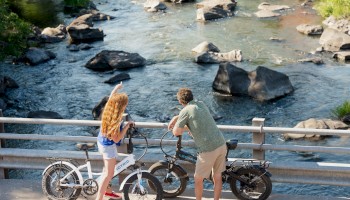 Two people with bicycles stand on a bridge, looking over a scenic river with rocks and greenery around it.