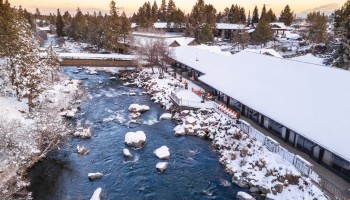 A snowy riverside scene with a river running through a town, snow-covered trees, and buildings along the riverbank under a cloudy sky.