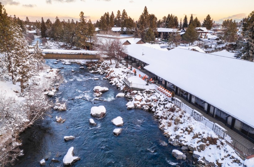 A snowy riverside scene with a river running through a town, snow-covered trees, and buildings along the riverbank under a cloudy sky.