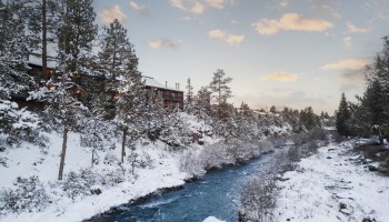Snowy landscape along the Deschutes River near Riverhouse Lodge, the best lodging location in Central Oregon, with snow-covered trees, a cozy cabin in the distance, and soft clouds overhead.