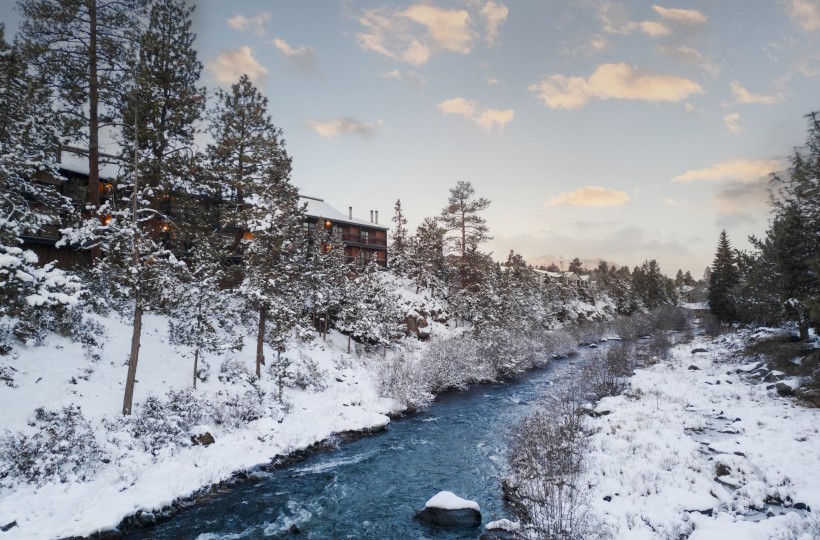 Snowy landscape along the Deschutes River near Riverhouse Lodge, the best lodging location in Central Oregon, with snow-covered trees, a cozy cabin in the distance, and soft clouds overhead.