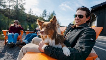 A person with sunglasses holds a brown and white dog on an orange chair outdoors at Riverhouse Lodge in Bend, Oregon, surrounded by trees and others sitting nearby.