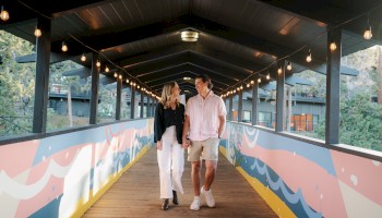 A couple holding hands and smiling as they walk across the covered pedestrian bridge with colorful murals and string lights at Riverhouse Lodge in Bend, Oregon.