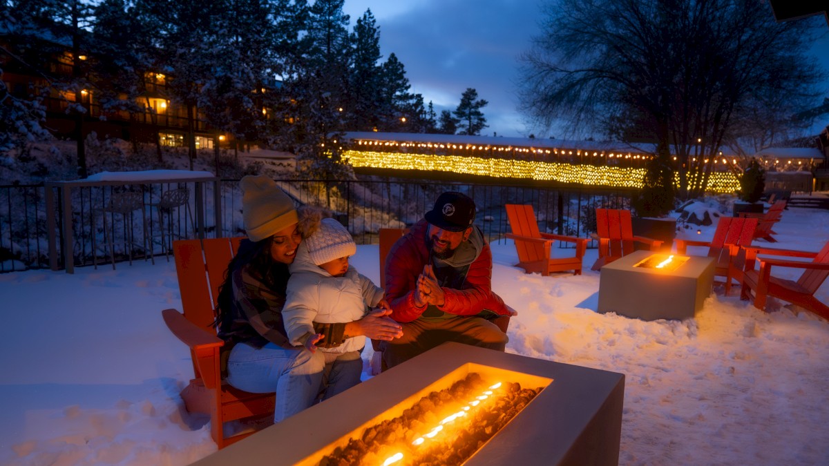 A family sits around a cozy outdoor fire pit, surrounded by snow and lit decorations, enjoying a winter evening in a serene setting.