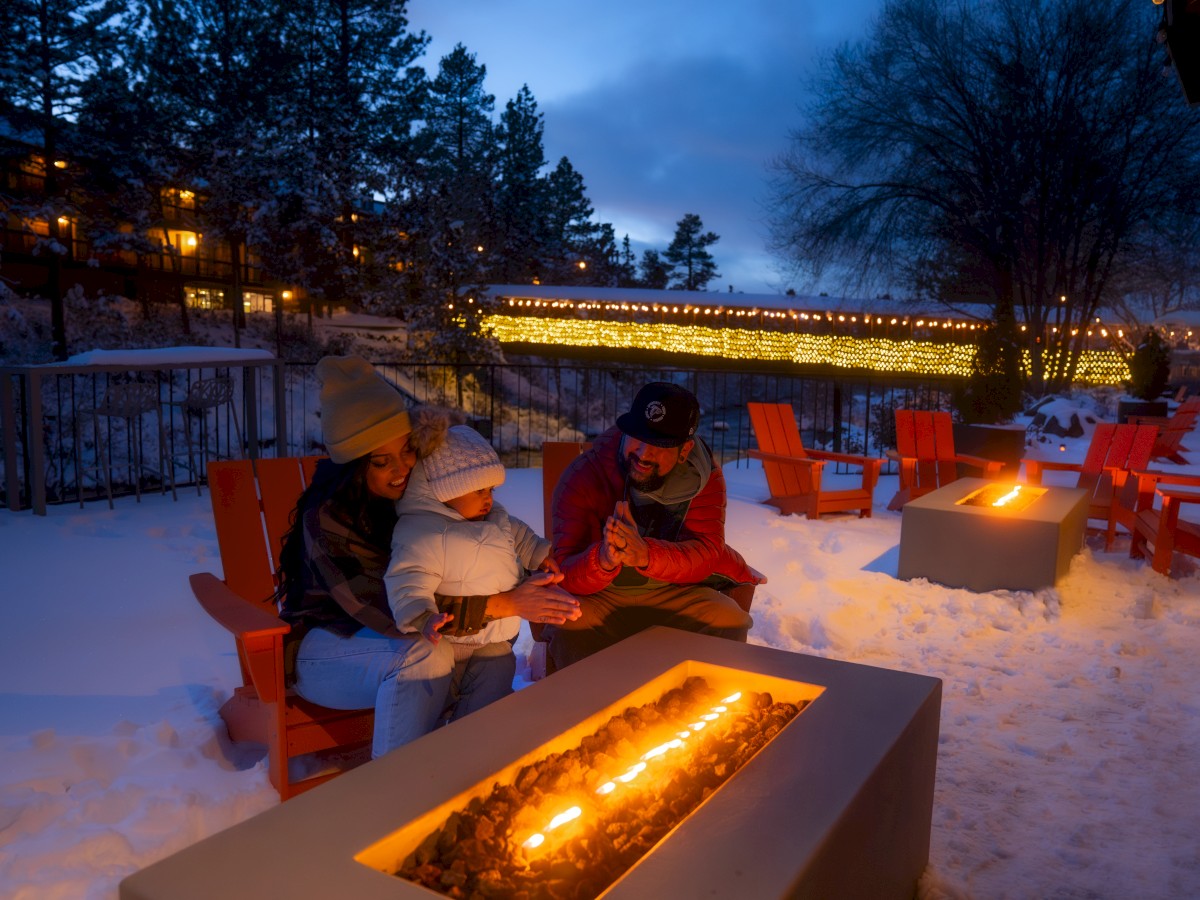 A family sits around a cozy outdoor fire pit, surrounded by snow and lit decorations, enjoying a winter evening in a serene setting.