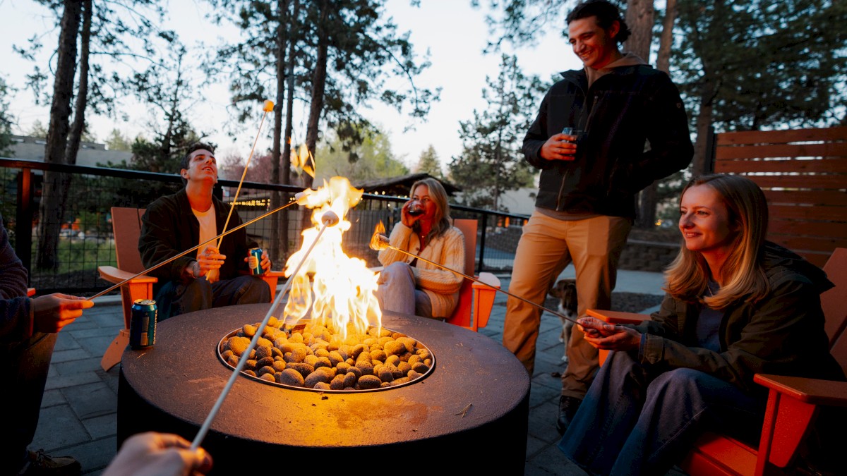 Guests roasting marshmallows around a glowing fire pit at Riverhouse Lodge in Bend, Oregon, surrounded by tall pine trees and cozy Adirondack chairs at dusk.