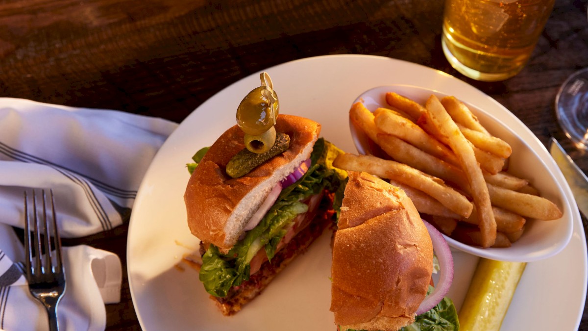 A plate with a cheeseburger, fries, and a pickle, alongside a beer and a flower vase, on a wooden table with a napkin and utensils at Currents at Riverhouse Lodge in Bend, Oregon which features views of the river for dining.