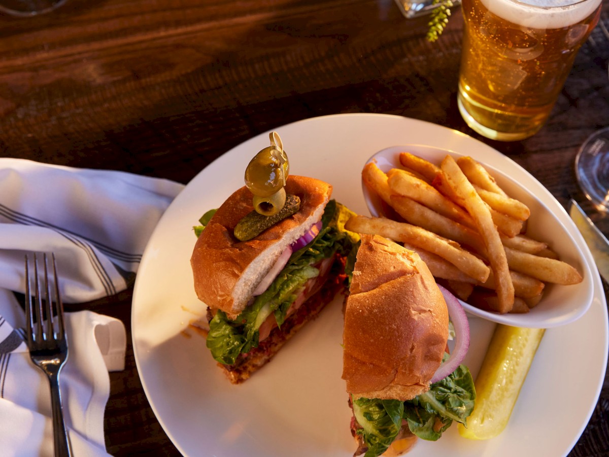 A plate with a sandwich, fries, and a pickle, alongside a beer and a flower vase, on a wooden table with a napkin and utensils.