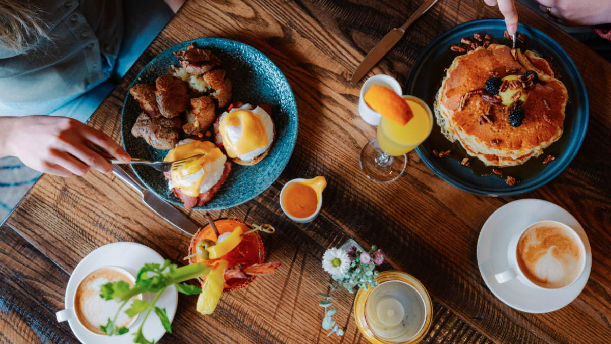 A brunch spread at Currents at the Riverhouse in Bend, Oregon with eggs Benedict, pancakes, drinks, and coffees on a wooden table, with hands reaching for food.