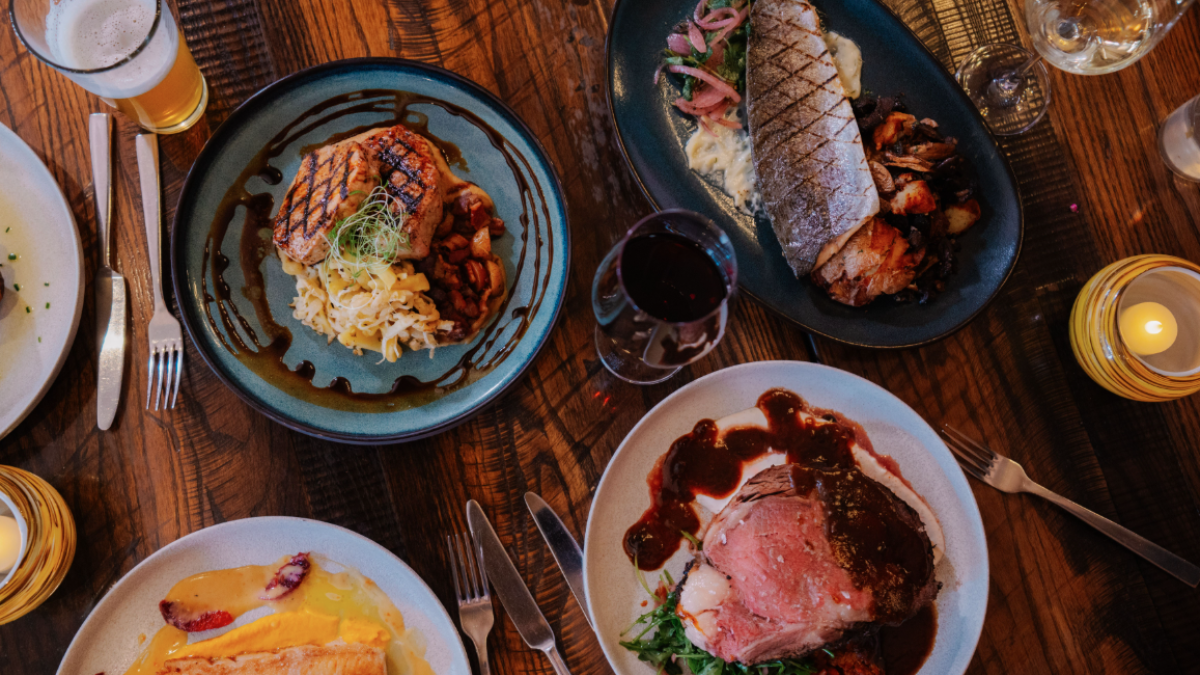 A table at Currents at the Riverhouse in Bend, Oregon with various gourmet dishes, including grilled fish, steak, and drinks, set on a wooden surface with utensils and a candle.
