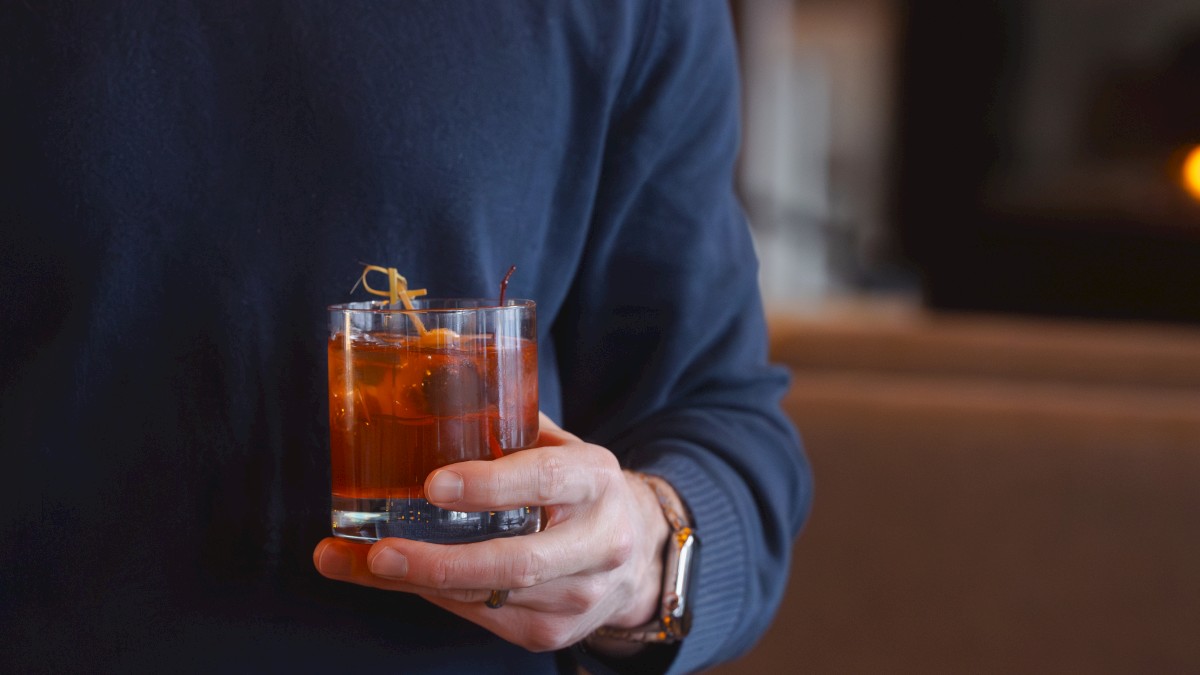 A person is holding a glass of a dark amber-colored drink with ice and a garnish at Currents at Riverhouse Lodge in Bend, Oregon.