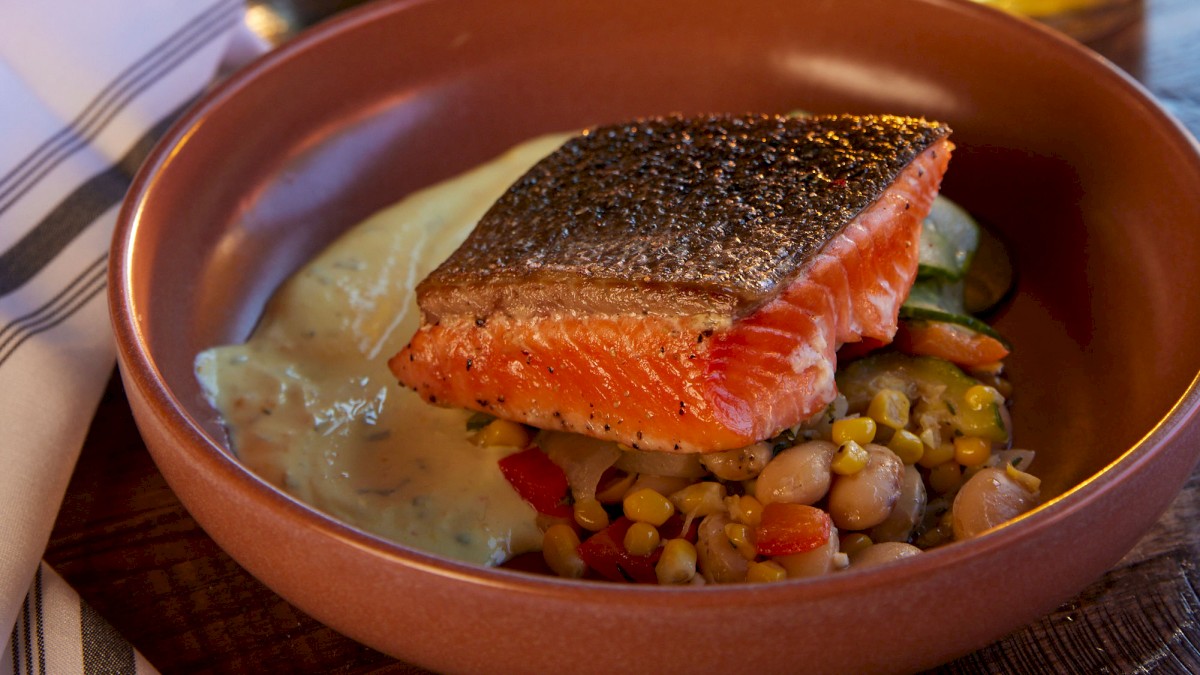 A meal featuring a salmon fillet over vegetables, a burger, a glass of beer, and a vase of flowers on a wooden table at Currents at Riverhouse Lodge, located in Bend, Oregon.